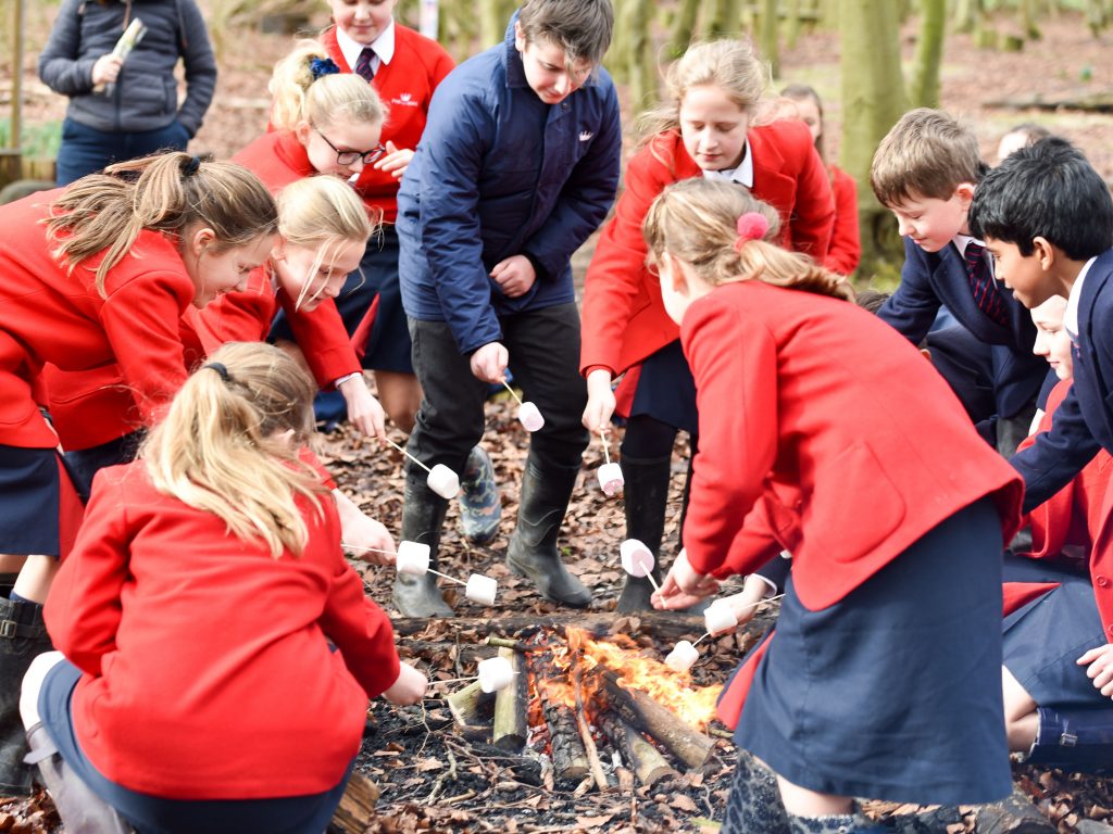 Kids in red blazers and navy coats stood around a campfire with marshmallows on sticks