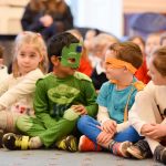 A group of kids sat on the floor of a classroom, all dressed up in different costumes, including superheroes and princesses.
