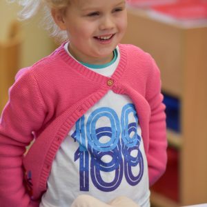 A young blonde girl wearing a pink cardigan, playing with a plastic baby doll.