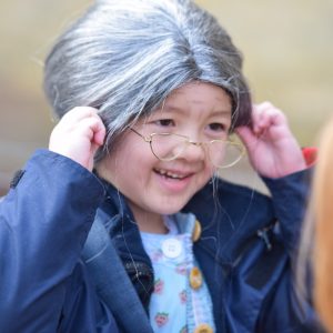 A small girl dressed up aas an old woman, with a grey wig and wire frame glasses.