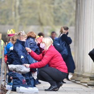 A mother in a red coat, wearing a facemask, zipping up their child's coat outside a school.